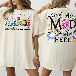 May include: Ivory-colored t-shirt featuring colorful "Name Wonderland 2026" text on the front. The back displays "We're All Mad Here" with Alice, the Cheshire Cat, and other Alice in Wonderland graphics. A whimsical design.