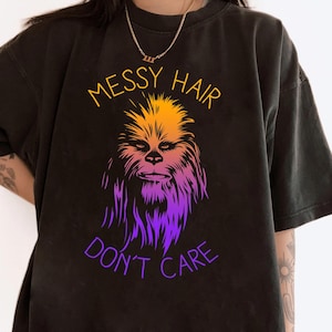 May include: Black t-shirt featuring a Wookiee-style character in a vibrant graphic. The text "MESSY HAIR DON'T CARE" is displayed in yellow and purple above and below the character. The t-shirt has a loose fit.