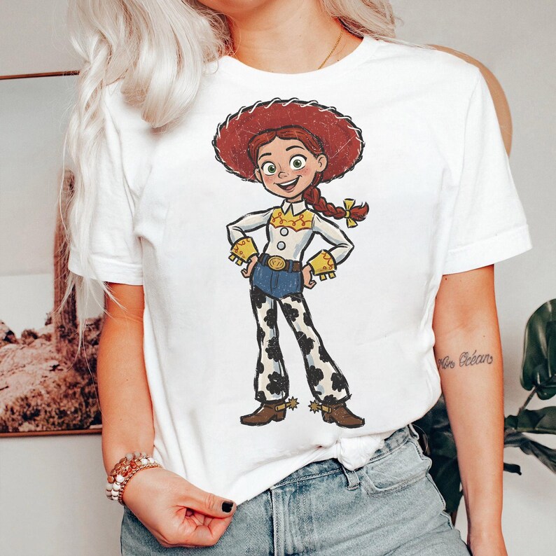 May include: A white short-sleeved t-shirt featuring a graphic print of a cartoon cowgirl character with red hair in a braid, a red hat, a white shirt with yellow accents, blue pants, and cow-print chaps, standing with hands on hips.