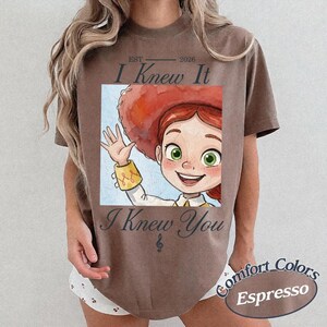 Peut inclure: Un t-shirt marron &agrave; manches courtes avec une impression graphique repr&eacute;sentant un personnage de dessin anim&eacute; aux cheveux roux et un grand chapeau rouge. Le texte indique EST 2026 I Knew It I Knew You avec une note de musique sous le texte.