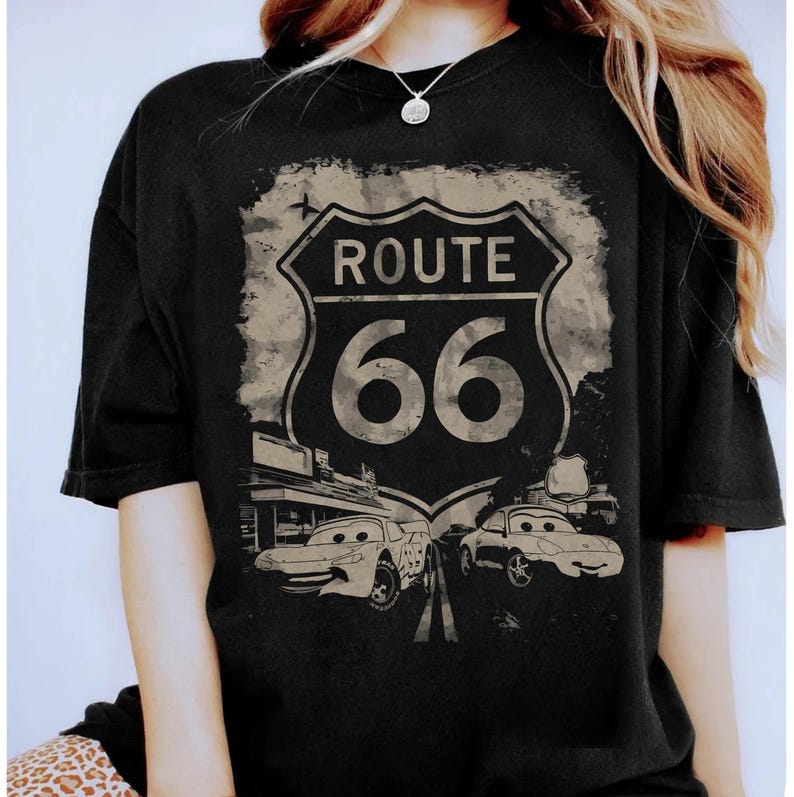 May include: A black short-sleeve t-shirt featuring a distressed graphic print of a Route 66 road sign. Below the sign, two cartoon-style cars with eyes on their windshields are depicted on a road, with a building structure visible on the left.