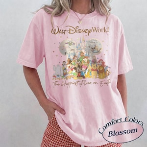 K&ouml;nnte beinhalten: Hellrosa T-Shirt mit einem Disney-Motiv, das Charaktere und den Text "Walt Disney World" und "The Happiest Place on Earth" zeigt. Das Shirt ist ein Comfort Colors Blossom T-Shirt.