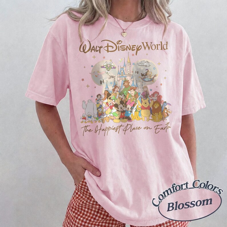 K&ouml;nnte beinhalten: Hellrosa T-Shirt mit einem Disney-Motiv, das Charaktere und den Text "Walt Disney World" und "The Happiest Place on Earth" zeigt. Das Shirt ist ein Comfort Colors Blossom T-Shirt.