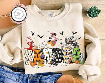 May include: Cream-colored sweatshirt featuring the name "SANDRA" in a Halloween-themed design with cartoon characters, pumpkins, bats, and spiderwebs. The letters are decorated with stripes, polka dots, and other patterns. A fun and festive top for the spooky season.