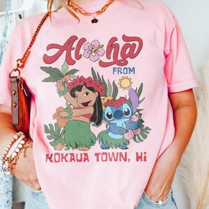 May include: Pink t-shirt with the words "Aloha From Kokaua Town, Hi" in a retro font. The shirt features a graphic of two cartoon characters in Hawaiian attire, surrounded by tropical flowers and foliage.