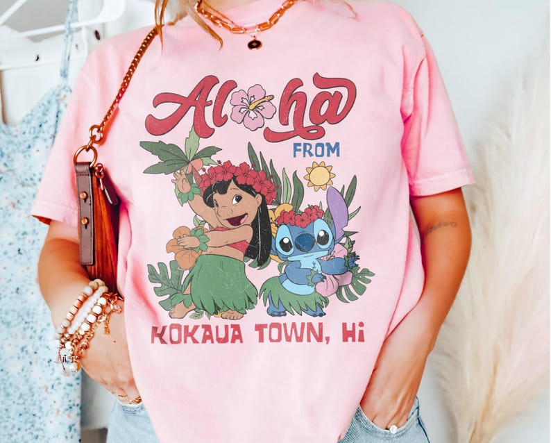 May include: Pink t-shirt with the words "Aloha From Kokaua Town, Hi" in a retro font. The shirt features a graphic of two cartoon characters in Hawaiian attire, surrounded by tropical flowers and foliage.