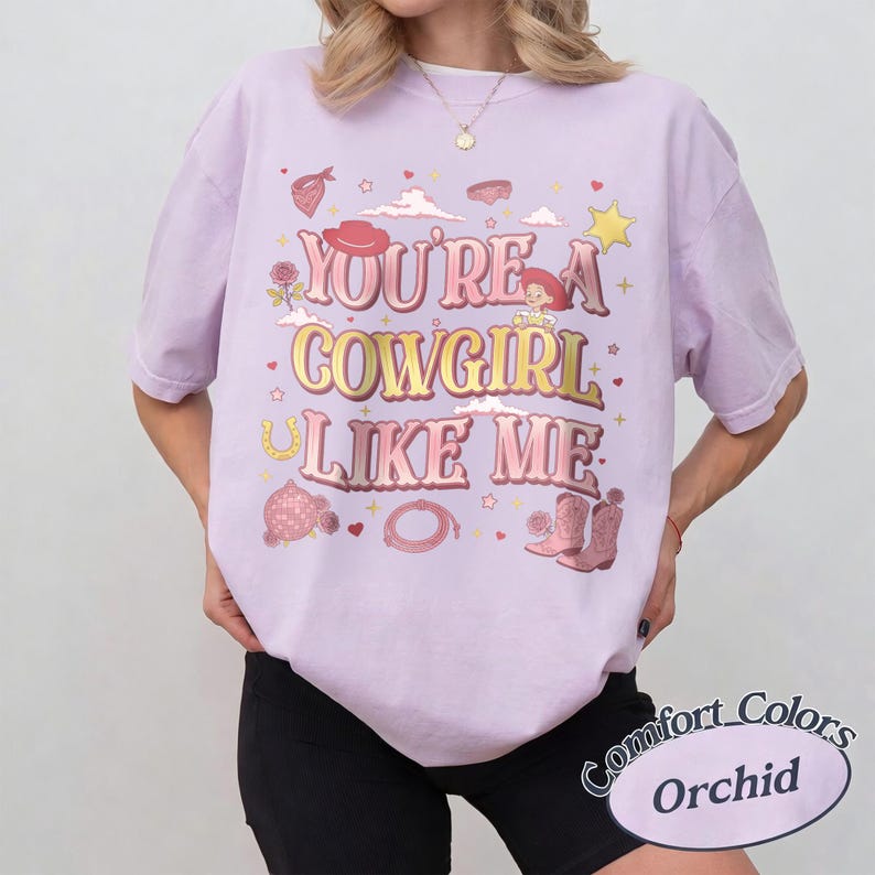 May include: A light purple short-sleeve t-shirt featuring the text YOU'RE A COWGIRL LIKE ME in pink and yellow gradient lettering. The graphic includes illustrations of a cowboy hat, a lasso, boots, a rose, a star, and a cartoon character.