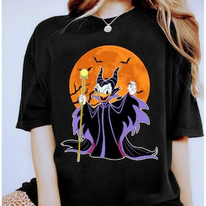 Op de afbeelding: Zwart T-shirt met Donald Duck als Maleficent, met staf, tegen een oranje maan met vleermuizen. De cape heeft paarse randen. Een Halloween-thema grafisch T-shirt.