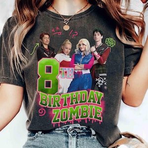 May include: Dark gray t-shirt featuring a colorful graphic design. The design includes the text "8th Birthday Zombie" in green and pink, along with images of characters. The shirt is a casual style.