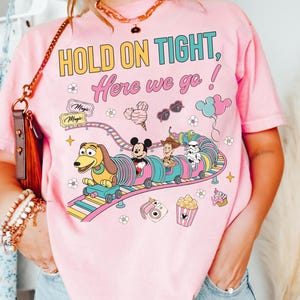 Op de afbeelding: Roze t-shirt met de tekst "HOLD ON TIGHT, Here we go!" en een kleurrijke afbeelding van een achtbaan met Disney-personages. Het ontwerp bevat Mickey Mouse, Woody en een Stormtrooper.