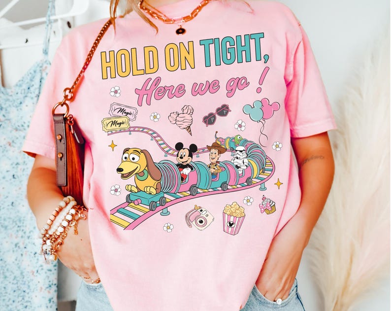 Op de afbeelding: Roze t-shirt met de tekst "HOLD ON TIGHT, Here we go!" en een kleurrijke afbeelding van een achtbaan met Disney-personages. Het ontwerp bevat Mickey Mouse, Woody en een Stormtrooper.