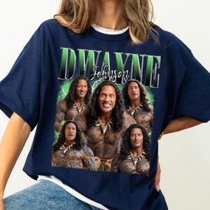 May include: Navy blue t-shirt featuring a graphic of Dwayne Johnson with the text "Dwayne Johnson" in a green and white design. The graphic includes multiple images of the actor. The shirt is a casual wear item.