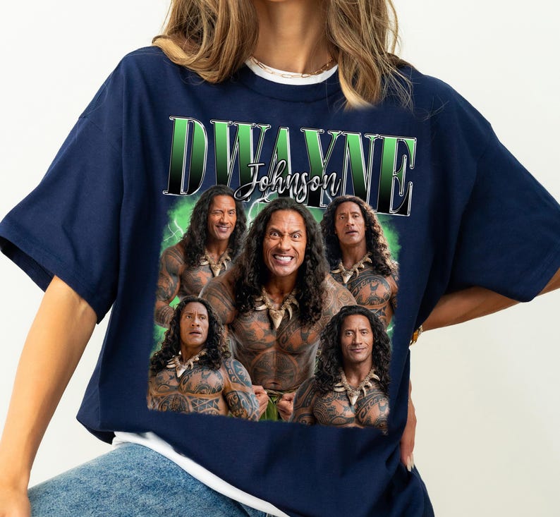 May include: Navy blue t-shirt featuring a graphic of Dwayne Johnson with the text "Dwayne Johnson" in a green and white design. The graphic includes multiple images of the actor. The shirt is a casual wear item.