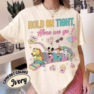 Op de afbeelding: Ivoor kleurig t-shirt met de tekst "HOLD ON TIGHT, Here we go!" en een kleurrijke afbeelding van Disney-figuren op een achtbaan. Het shirt bevat ook de woorden "COMFORT COLORS".