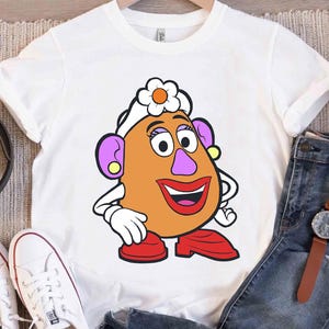 May include: White t-shirt featuring a cartoon character with an orange face, purple ears, and a red mouth. The character has a white flower on its head and is wearing red shoes. The shirt is displayed with jeans and white sneakers.
