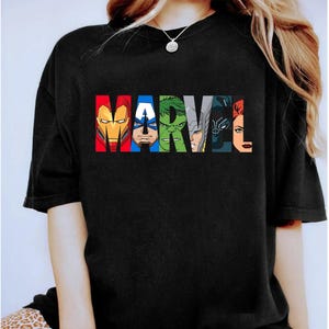 May include: Black t-shirt featuring the word "MARVEL" in colorful block letters. Each letter is filled with a different superhero's face: Iron Man, Captain America, Hulk, Thor, Black Panther, and Black Widow. A casual, graphic tee.