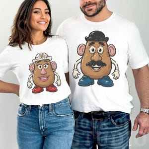 May include: Two white short-sleeved t-shirts. The left shirt features a graphic of a potato character wearing a white hat with a flower, gold hoop earrings, and red shoes. The right shirt features a potato character with a black bowler hat and blue shoes.