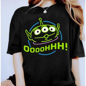 Puede incluir: Camiseta negra con un diseño de extraterrestre verde de dibujos animados. El extraterrestre tiene tres ojos y orejas grandes, con el texto "OoooHH!" en verde y azul. El extraterrestre está dentro de un círculo azul con un contorno rosa. La camiseta es de estilo casual.