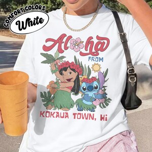 May include: White t-shirt with a colorful graphic featuring the words "Aloha From Kokaua Town, Hi" and illustrations of two cartoon characters. The shirt has a round neckline and short sleeves.