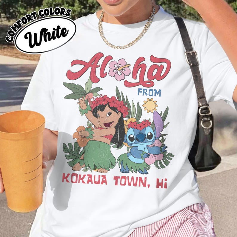 May include: White t-shirt with a colorful graphic featuring the words "Aloha From Kokaua Town, Hi" and illustrations of two cartoon characters. The shirt has a round neckline and short sleeves.