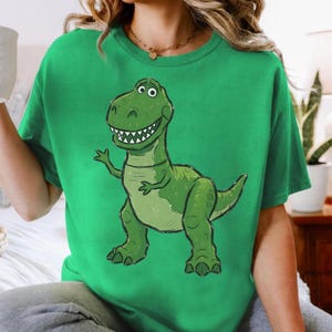 May include: A bright green short-sleeved t-shirt featuring a large, centered graphic illustration of a smiling green Tyrannosaurus Rex dinosaur with a light-colored belly, sharp teeth, and small arms, rendered in a cartoon style on the front.