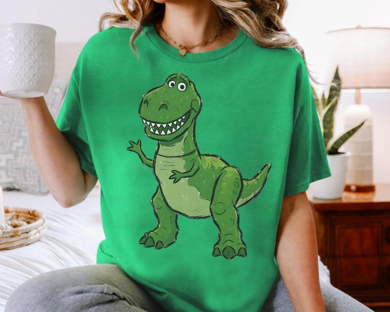 May include: A bright green short-sleeved t-shirt featuring a large, centered graphic illustration of a smiling green Tyrannosaurus Rex dinosaur with a light-colored belly, sharp teeth, and small arms, rendered in a cartoon style on the front.