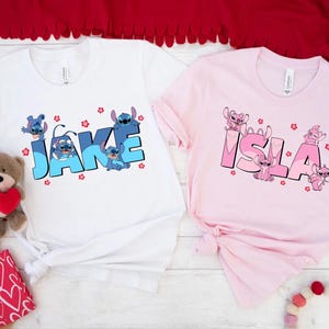 May include: Two t-shirts, one white and one pink, each featuring a cartoon design. The white shirt displays the name "Jake" in blue block letters with Stitch characters and red flower accents. The pink shirt has the name "Isla" in pink with Stitch characters.