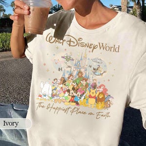 K&ouml;nnte beinhalten: Elfenbeinfarbenes T-Shirt mit dem Schriftzug "Walt Disney World" in einer stilisierten Schriftart. Darunter befindet sich eine farbenfrohe Grafik von Disney-Figuren und einem Schloss. Der Satz "The Happiest Place on Earth" ist unter der Grafik aufgedruckt.