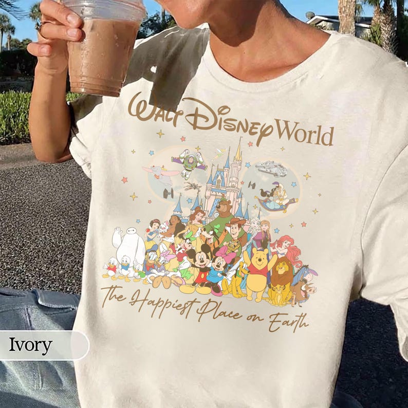 K&ouml;nnte beinhalten: Elfenbeinfarbenes T-Shirt mit dem Schriftzug "Walt Disney World" in einer stilisierten Schriftart. Darunter befindet sich eine farbenfrohe Grafik von Disney-Figuren und einem Schloss. Der Satz "The Happiest Place on Earth" ist unter der Grafik aufgedruckt.