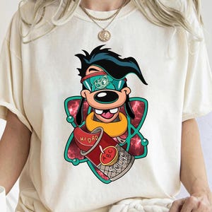 May include: Cream-colored t-shirt featuring a cartoon character with black hair, sunglasses, and a yellow collar. The character holds a red can of soup with the text "Hi Dad" and "ABC Soup". The graphic is set against a red and green background.
