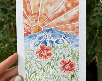 Puede incluir: Pintura a la acuarela de un paisaje montañoso con una puesta de sol y flores silvestres. La obra de arte mide 12,7 cm x 17,8 cm y presenta una vibrante paleta de colores naranja y azul. El texto "MT. BACHELOR WILDFLOWERS 2023" está escrito en la parte inferior.