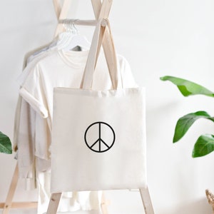 Könnte beinhalten: Eine weiße Canvas-Tragetasche mit einem schwarzen Peace-Zeichen auf der Vorderseite. Die Tasche hängt an einem Holzgestell mit anderer Kleidung.