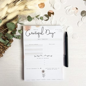 Könnte beinhalten: Ein weißer Notizblock mit einem Aquarellhintergrund und dem Text "have a Grateful Day". Der Notizblock ist in Abschnitte für "Mein Tagesziel", "Prioritäten", "To Do's & Termine" und "Notizen" unterteilt. Es gibt auch einen Abschnitt für "Ich bin dankbar für..." und "Selbstpflege - das tue ich heute für mich".