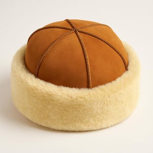 May include: A brown hat with a rounded crown and a cream-colored, fluffy brim. The crown is divided into sections by stitched brown lines. The hat appears warm and is likely made of a soft material, suitable for cold weather.