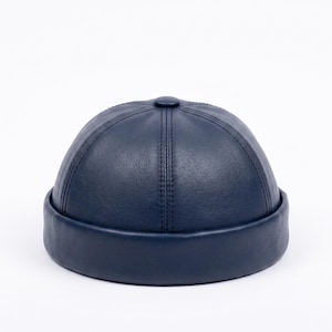 May include: A navy blue docker cap featuring a smooth, leather-like texture. The hat is constructed with six panels meeting at a central button on the crown, finished with a wide, rolled brim that encircles the base of the dome-shaped headwear.