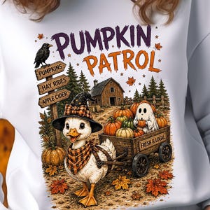 May include: A white sweatshirt featuring a graphic of a duck wearing a plaid witch hat and scarf, pulling a wooden wagon filled with pumpkins and a ghost. The design includes a signpost labeled PUMPKINS, HAY RIDES, and APPLE CIDER, with PUMPKIN PATROL text.