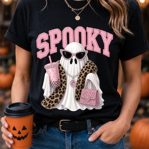 Puede incluir: Una camiseta negra con el gráfico de un fantasma blanco que lleva gafas de sol de ojo de gato rosas, una bufanda de leopardo y un collar de cristal. El fantasma sostiene una bebida boba rosa y un bolso rosa. La palabra SPOOKY está impresa en rosa sobre el fantasma.