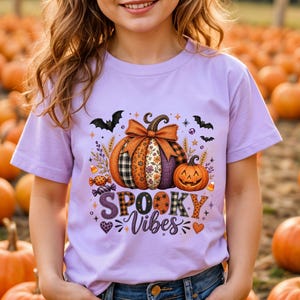 May include: A light purple t-shirt featuring a graphic print of two pumpkins, one large with a bow and one small jack-o'-lantern, surrounded by bats, candy, and wheat stalks. The text "SPOOKY Vibes" is printed in purple and orange patterned letters.