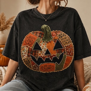 May include: A charcoal gray short-sleeve t-shirt featuring a large, central pumpkin graphic made of various fabric patches with visible stitching. The pumpkin includes the text "HELLO FALL," "SPOOKY SEASON," and "STAY COZY, BE SPOOKY."