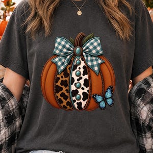 Puede incluir: Una camiseta gris carbón con el gráfico de una calabaza naranja con un lazo de cuadros azul y blanco, un panel central con estampado de vaca y piedras turquesas, y una mariposa azul posada en el lateral de la ilustración de la calabaza.