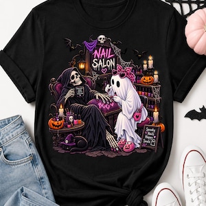 May include: A black t-shirt featuring a graphic print of a skeleton and a ghost in a nail salon. The skeleton sits in a chair while the ghost paints its nails. The design includes text reading NAIL SALON, BOO, and Spooky Nails Happy Souls.