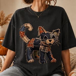 以下が含まれることがあります： チャコールグレーの半袖Tシャツで、様々な布のパッチを組み合わせて作られた猫の大きなグラフィックが特徴です。猫のデザインには、デニム、チェック柄、無地の布地が含まれており、ほつれた端やステッチのディテールがはっきりと見えています。