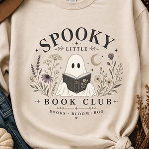 May include: A cream-colored crewneck sweatshirt featuring a graphic of a ghost reading a book, surrounded by botanical illustrations, a crescent moon, and stars. The text reads SPOOKY LITTLE BOOK CLUB and BOOKS BLOOM BOO in black lettering.