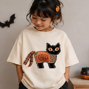 以下が含まれることがあります： オレンジ色の目をした黒猫のグラフィックが描かれたクリーム色の半袖Tシャツ。猫の体はパッチワーク風で、HELLO FALLと書かれたオレンジ色の部分、ヒョウ柄の部分、チェック柄の尻尾があり、ステッチのディテールが施されています。