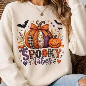 May include: A cream-colored crewneck sweatshirt featuring a graphic print of two pumpkins, one with a large orange bow and patterned segments, and the other a carved jack-o'-lantern. The text reads SPOOKY Vibes, surrounded by bats, candy, and stars.