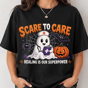 May include: A black t-shirt featuring a graphic of a ghost dressed as a nurse with a stethoscope and clipboard. The design includes a spider, a bat, and a jack-o'-lantern, with the text "SCARE TO CARE" and "HEALING IS OUR SUPERPOWER" in orange.