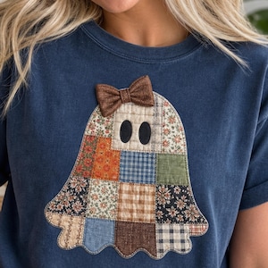 May include: A navy blue t-shirt featuring a large ghost-shaped appliqué made from various patchwork fabric squares in floral, plaid, and solid patterns. The ghost has two black oval eyes and a small brown textured bow attached at the top.
