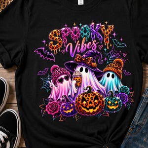 May include: A black t-shirt featuring a colorful graphic with the text "SPOOKY Vibes" in a dripping font. The design includes three ghosts wearing leopard print hats, a witch hat, and sunglasses, surrounded by pumpkins, roses, and bats.