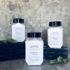 May include: Three white plastic bottles with black lids containing carpet freshener. The bottles are labeled "White Bark Cottage" and "Magnolia + Peony". The bottles are sitting on a wooden surface.