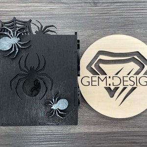 May include: Black wooden box with a spider web design and three silver spiders. The box is next to a round wooden sign with a diamond shape and the text "GEM:DESIGN".